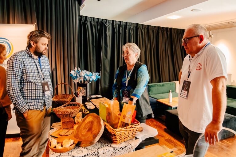 Trzy osoby rozmawiają przy stoisku z regionalnymi produktami w Mińsku Mazowieckim. Na stole znajdują się kosze z chlebem, drewniane naczynia i torby z lokalnymi wyrobami. W tle widoczne są ciemne zasłony i elementy wystroju sali.