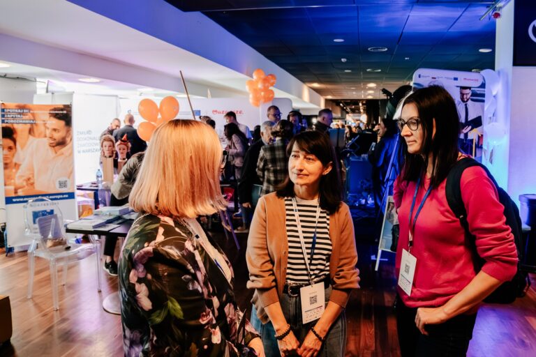 Trzy kobiety rozmawiają podczas wydarzenia edukacyjnego w hali wystawienniczej. W tle widać stoiska informacyjne, plakaty, pomarańczowe balony i uczestników targów.