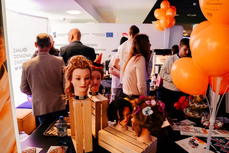 Stoisko edukacyjne prezentujące fryzjerskie główki treningowe z różnymi fryzurami i ozdobami. W tle osoby rozmawiające przy ekspozycji z pomarańczowymi balonami i materiałami promocyjnymi Zakładu Doskonalenia Zawodowego.