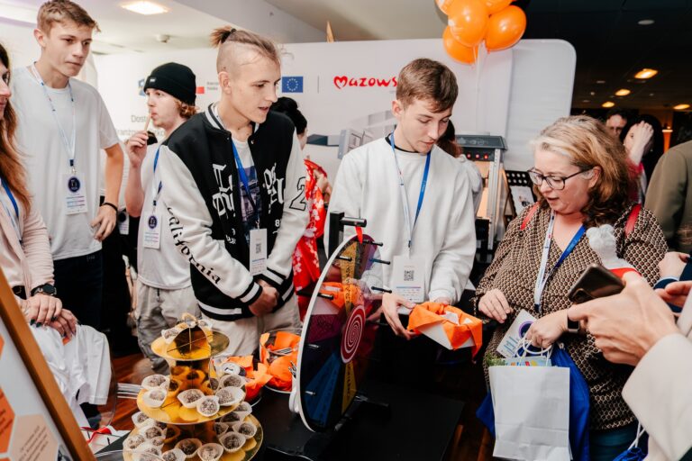 Grupa młodych osób i kobieta biorą udział w wydarzeniu edukacyjnym przy stoisku z kołem fortuny. Uczestnicy trzymają torby z materiałami promocyjnymi, a w tle widoczne są logotypy Mazowsza i Unii Europejskiej oraz balony w kolorze pomarańczowym.