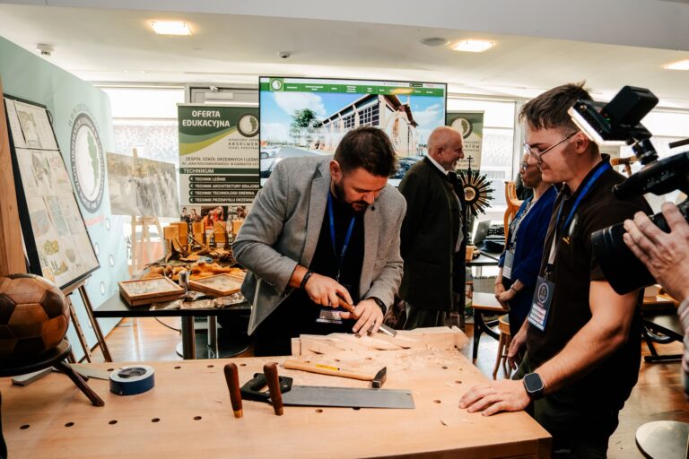 Mężczyzna w szarej marynarce wykonuje rzeźbienie w drewnie na warsztacie stolarskim podczas prezentacji edukacyjnej. Obok niego stoją uczestnicy wydarzenia obserwujący pracę, a w tle widoczne są plakaty i materiały promujące szkołę leśną i drzewną.