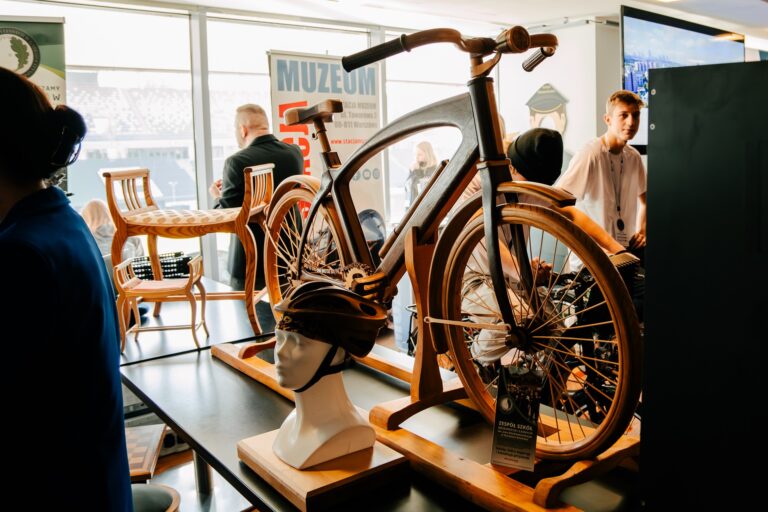 Na zdjęciu znajduje się ekspozycja z drewnianym rowerem ustawionym na stole wystawowym. Obok umieszczono manekinową głowę z kaskiem rowerowym oraz krzesła o drewnianej konstrukcji. W tle widoczny jest baner Stacji Muzeum oraz osoby uczestniczące w wydarzeniu.