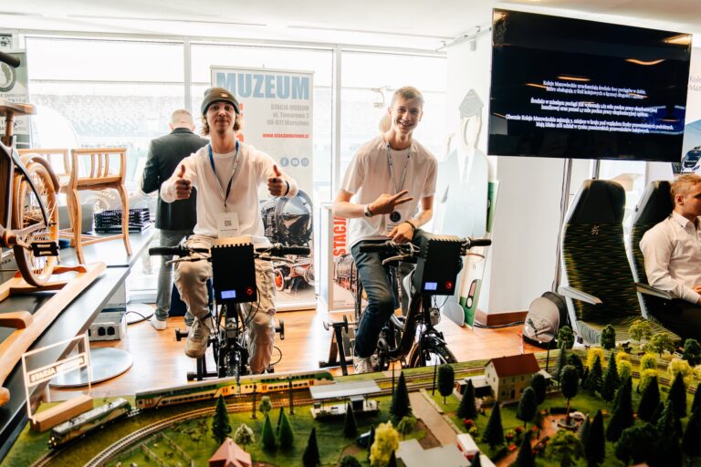 Na zdjęciu dwóch młodych mężczyzn w białych koszulkach siedzi na rowerach stacjonarnych, uśmiecha się i pokazuje gesty „OK” oraz „pokój”. Przed nimi znajduje się makieta kolejowa z miniaturowymi pociągami i budynkami. W tle widać baner Stacji Muzeum oraz monitor z prezentacją.