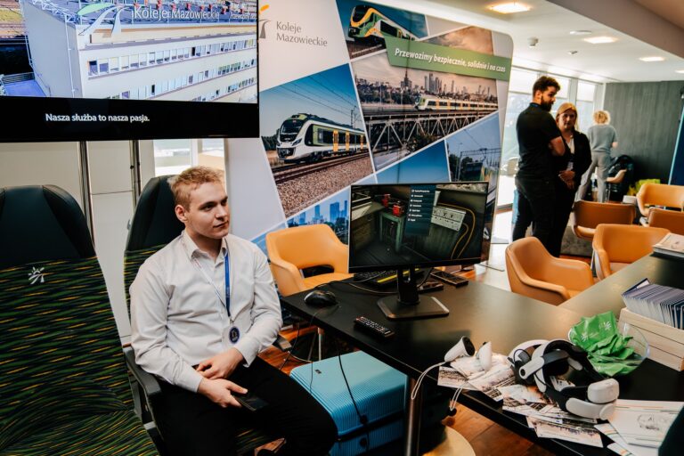 Na zdjęciu stoisko Kolei Mazowieckich podczas wydarzenia. Mężczyzna w białej koszuli siedzi na fotelu pasażerskim obok stanowiska z monitorem prezentującym symulację kolejową. Na stole leżą broszury, okulary VR i mysz komputerowa. W tle widoczne są banery promujące bezpieczeństwo i punktualność przewoźnika oraz kilku uczestników rozmowy.