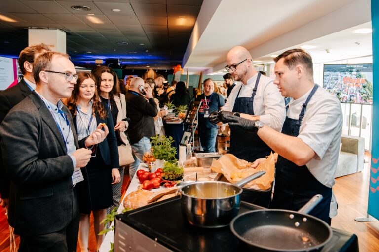 Na zdjęciu kucharze w białych koszulach i granatowych fartuchach przygotowują potrawy przy stanowisku kulinarnym podczas wydarzenia. Na stole widać świeże zioła, chleb i warzywa. Naprzeciwko nich stoi grupa uczestników degustujących potrawy i rozmawiających z kucharzami. W tle znajdują się inne stoiska i odwiedzający.