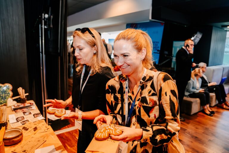Na zdjęciu dwie kobiety stoją przy stole wystawowym i trzymają w dłoniach ozdobne pierniki. Obie uśmiechają się i oglądają przygotowane wypieki. Na stole leżą broszury, karty informacyjne i gliniane naczynia. W tle widać uczestników wydarzenia siedzących na sofach.
