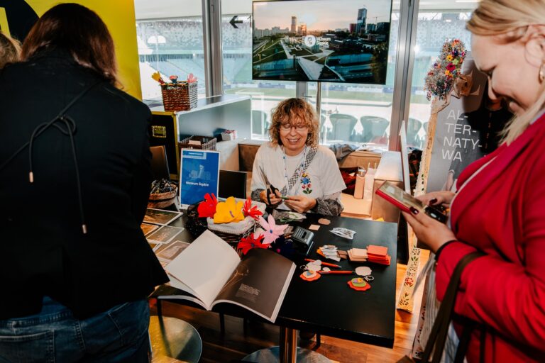 Na zdjęciu stoisko Muzeum Śląskiego prezentujące projekt „ETNO Alternatywa”. Kobieta siedząca przy stole wykonuje kolorowe kwiaty z papieru, wokół niej znajdują się zdjęcia, broszury, książka i materiały plastyczne. W tle widać ekran z logo muzeum oraz osoby odwiedzające stoisko.