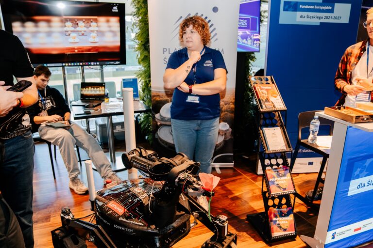 Na zdjęciu stoisko województwa śląskiego prezentujące projekty Politechniki Śląskiej w ramach Funduszy Europejskich 2021–2027. Na pierwszym planie znajduje się robot mobilny z elementami technologicznymi i ozdobnym czerwonym kwiatem. Za nim stoją uczestnicy wydarzenia, a w tle widoczne są banery promujące Platformę Śląską i publikacje uczelni.