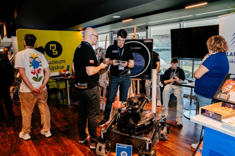 Na zdjęciu stoisko przedstawiające projekty Politechniki Śląskiej podczas wydarzenia. Na pierwszym planie znajduje się robot mobilny z elementami mechanicznymi i ozdobnym czerwonym kwiatem. Wokół stoją uczestnicy wydarzenia — niektórzy rozmawiają, inni korzystają z tabletów lub komputerów. W tle widoczne są banery Politechniki Śląskiej i Muzeum Śląskiego.