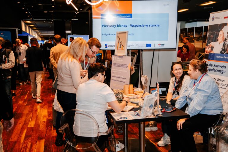 Na zdjęciu kilka osób rozmawia przy stoisku informacyjnym dotyczącym programu „Pierwszy biznes – Wsparcie w starcie”, realizowanego w ramach Funduszy Europejskich. Na stole leżą materiały promocyjne, a w tle widoczne są banery i ekran z prezentacją. Scena ma miejsce w przestrzeni wystawienniczej województwa dolnośląskiego.