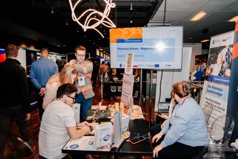 Na zdjęciu kilka osób rozmawia przy stoisku informacyjnym dotyczącym programu „Pierwszy biznes – Wsparcie w starcie”, realizowanego w ramach Funduszy Europejskich. Na stole leżą materiały promocyjne, a w tle widoczne są banery i ekran z prezentacją. Scena ma miejsce w przestrzeni wystawienniczej województwa dolnośląskiego.