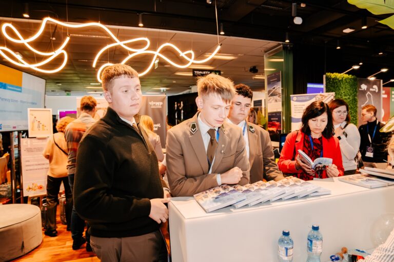 Na zdjęciu widać grupę osób stojących przy stoisku informacyjnym podczas wydarzenia. Kilku młodych mężczyzn w mundurach leśnych oraz kobieta w czerwonym żakiecie oglądają materiały promocyjne ułożone na blacie. W tle widoczne są inne stoiska wystawiennicze i uczestnicy targów.