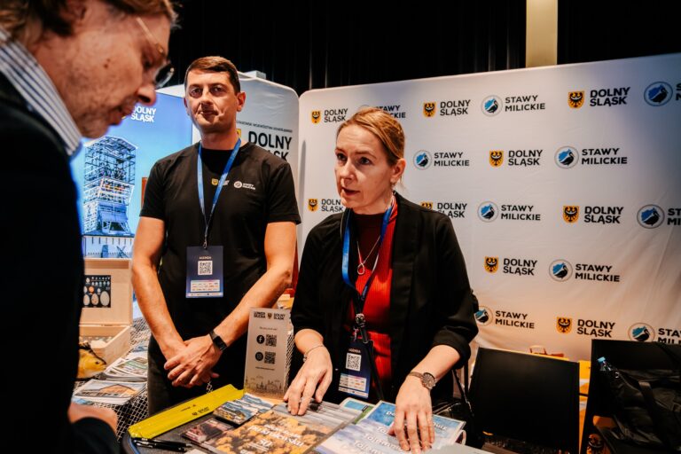 Na zdjęciu przedstawiciele województwa dolnośląskiego i Stawów Milickich rozmawiają z uczestnikiem wydarzenia przy stoisku promocyjnym. Na stole znajdują się foldery i materiały informacyjne dotyczące regionu, a w tle widoczne są banery z logotypami Dolnego Śląska i Stawów Milickich.