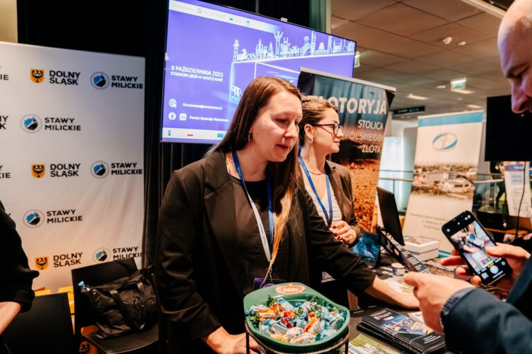 Dwie kobiety stoją przy stoisku promocyjnym województwa dolnośląskiego i Stawów Milickich, rozmawiając z odwiedzającymi. Na stole przed nimi leżą materiały informacyjne i miska ze słodyczami, a w tle widać banery i ekrany z informacjami o Forum Rozwoju Mazowsza.