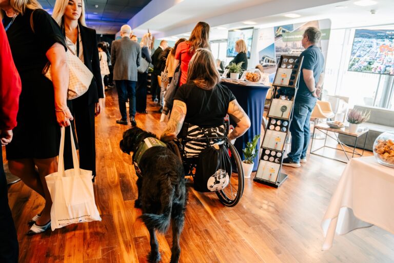 Uczestniczka wydarzenia poruszająca się na wózku z psem asystującym podczas 15. Forum Rozwoju Mazowsza – przykład integracji i dostępności przestrzeni targowej dla wszystkich odwiedzających.