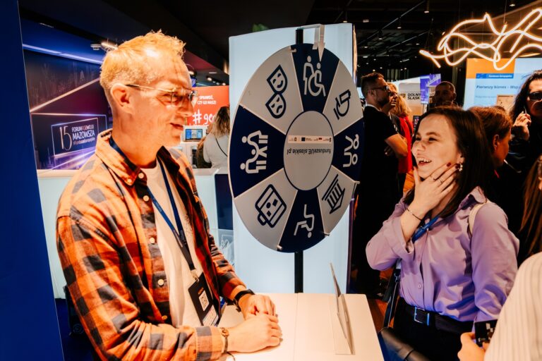 Uczestnicy 15. Forum Rozwoju Mazowsza biorą udział w interaktywnej zabawie przy kole fortuny w strefie Funduszy Europejskich – rozmowa w przyjaznej atmosferze przyciąga uwagę odwiedzających.