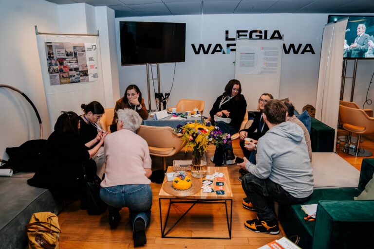 Uczestnicy 15. Forum Rozwoju Mazowsza podczas rozmowy w strefie warsztatowej – grupa osób dyskutuje przy stoliku w przyjaznej atmosferze, w otoczeniu materiałów informacyjnych i kwiatów.