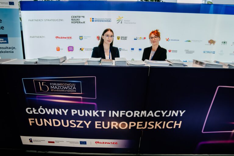 Dwie kobiety obsługujące główny punkt informacyjny Funduszy Europejskich podczas 15. Forum Rozwoju Mazowsza.