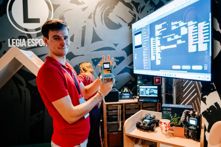 Uczestnik 15. Forum Rozwoju Mazowsza prezentuje urządzenie elektroniczne z wyświetlaczem LED na stoisku poświęconym nowym technologiom i programowaniu. W tle widoczny jest monitor z interfejsem graficznym do programowania oraz roboty edukacyjne wykorzystywane do demonstracji zasad automatyki i kodowania.