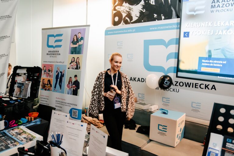 Stoisko Akademii Mazowieckiej w Płocku podczas 15. Forum Rozwoju Mazowsza. Na banerach widoczne logo uczelni oraz informacje o kierunkach studiów, w tym bezpieczeństwie wewnętrznym, ekonomii i kierunku lekarskim. Przy stoisku stoi przedstawicielka uczelni z identyfikatorem, a wokół rozmieszczone są materiały promocyjne, ulotki i gadżety z logo Akademii.