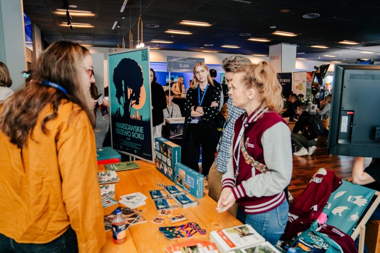 Stoisko m.st. Warszawy podczas 15. Forum Rozwoju Mazowsza, promujące inicjatywy ekologiczne miasta. Na środku widoczny plakat „Warszawskie Drzewo Roku”, a na stole rozłożone są broszury, ulotki i gry edukacyjne związane z ochroną środowiska. Kilka osób rozmawia przy stoisku, a w tle widać inne ekspozycje targowe i uczestników wydarzenia.