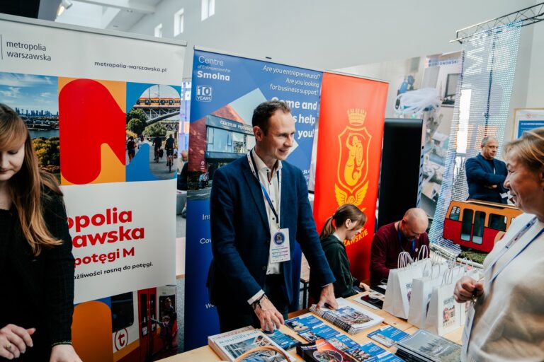 Stoisko Tramwajów Warszawskich i Centrum Przedsiębiorczości Smolna podczas 15. Forum Rozwoju Mazowsza. Za stołem siedzi dwoje przedstawicieli z identyfikatorami, obok nich stoi mężczyzna rozmawiający z odwiedzającymi. Na tle widoczne są kolorowe roll-upy z logotypami instytucji. Na stole rozłożone są broszury, ulotki i gadżety promujące Warszawę oraz dzielnicę Pragę.