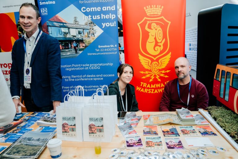 Stoisko Tramwajów Warszawskich i Centrum Przedsiębiorczości Smolna podczas 15. Forum Rozwoju Mazowsza. Za stołem siedzi dwoje przedstawicieli z identyfikatorami, obok nich stoi mężczyzna rozmawiający z odwiedzającymi. Na tle widoczne są kolorowe roll-upy z logotypami instytucji. Na stole rozłożone są broszury, ulotki i gadżety promujące Warszawę oraz dzielnicę Pragę.