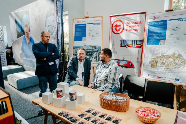 Stoisko Warszawskiego Transportu Publicznego podczas 15. Forum Rozwoju Mazowsza. Przy stole siedzi trzech mężczyzn prowadzących rozmowę. W tle widoczne są plansze informacyjne o projektach „Parkuj i Jedź” oraz wizualizacje inwestycji przy stacjach metra. Na stole leżą ulotki, kubki z logo komunikacji miejskiej i koszyk z gadżetami.