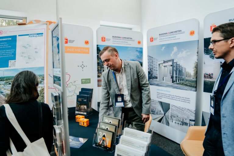 Stoisko Urzędu m.st. Warszawy podczas 15. Forum Rozwoju Mazowsza, prezentujące inwestycje miejskie. Na planszach widoczne są wizualizacje i mapy dotyczące budowy II linii metra oraz planowanej linii M3. Przy stoliku stoją przedstawiciele miasta rozmawiający z odwiedzającymi, a na blacie znajdują się broszury i materiały informacyjne.