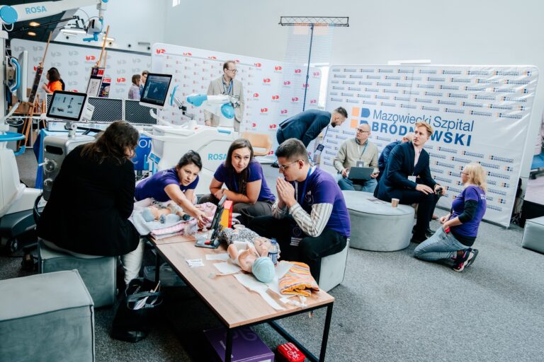 Stoisko Mazowieckiego Szpitala Bródnowskiego podczas 15. Forum Rozwoju Mazowsza. Na pierwszym planie zespół w fioletowych koszulkach prowadzi pokaz udzielania pierwszej pomocy niemowlętom z użyciem manekinów szkoleniowych. W tle widoczne logo „Mazowiecki Szpital Bródnowski” oraz nowoczesny sprzęt medyczny, w tym robot chirurgiczny ROSA.