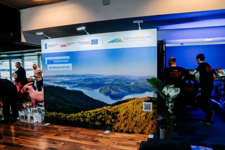 Stoisko Województwa Podkarpackiego podczas 15. Forum Rozwoju Mazowsza. Na ściance widnieje duża fotografia Jeziora Solińskiego z napisem „Fundusze Europejskie dla Podkarpacia 2021–2027” oraz logo regionu „Podkarpackie – przestrzeń otwarta”. Po prawej stronie widać uczestników korzystających z symulatorów jazdy oznaczonych logo „ESA”.