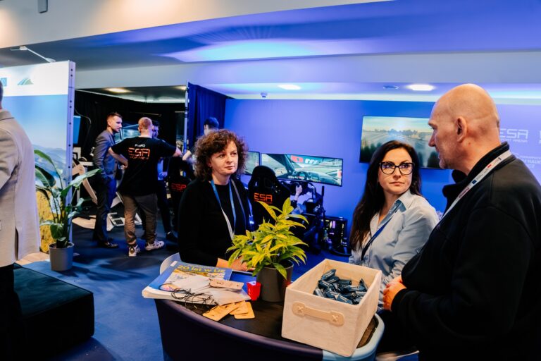 Stoisko Województwa Podkarpackiego podczas 15. Forum Rozwoju Mazowsza. Na pierwszym planie trzy osoby rozmawiają przy stoliku z materiałami promocyjnymi i rośliną, a w tle widać stanowiska symulatorów jazdy oznaczone logo „ESA”. Oświetlenie w tonacji niebieskiej tworzy nowoczesny, technologiczny klimat.
