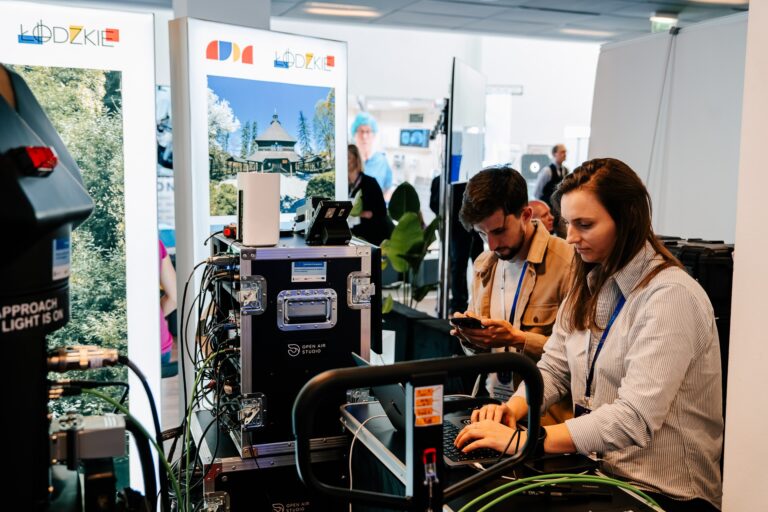 Stoisko Województwa Łódzkiego podczas 15. Forum Rozwoju Mazowsza. Dwoje pracowników obsługuje sprzęt techniczny i komputerowy oznaczony logo „Open Air Studio”, przygotowując transmisję lub nagranie. W tle widoczne są plansze promujące region łódzki z krajobrazami i architekturą.