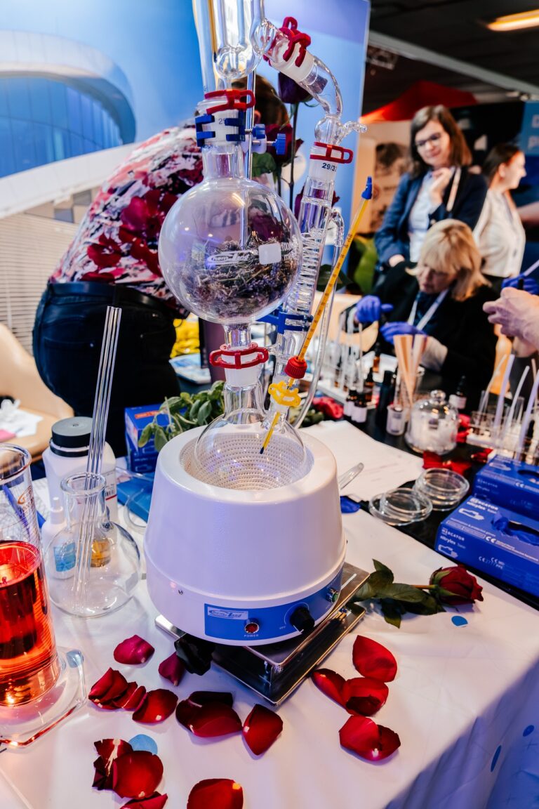 Stanowisko pokazowe z aparaturą laboratoryjną do destylacji olejków eterycznych podczas 15. Forum Rozwoju Mazowsza. Na stole stoi kolba z chłodnicą i rurkami wypełniona suszem roślinnym, otoczona płatkami róż i probówkami. W tle widać uczestniczki warsztatów zapachowych pracujące z próbkami aromatów.