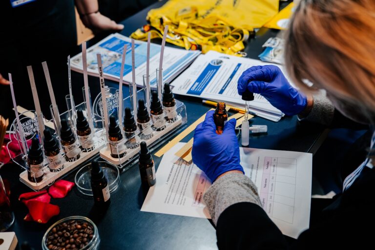 Osoba w niebieskich rękawiczkach laboratoryjnych przygotowuje próbkę zapachową przy stanowisku z buteleczkami olejków eterycznych podczas 15. Forum Rozwoju Mazowsza. Na stole widać paski testowe, płatki róż i ziarna kawy, używane do neutralizacji zapachów.