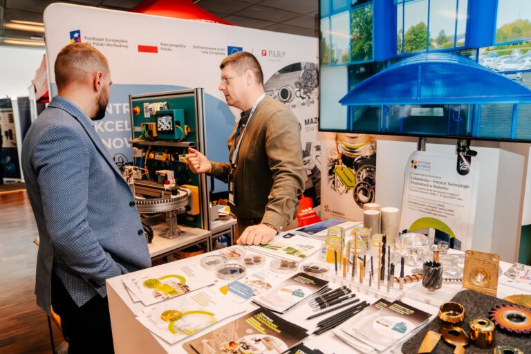 Stoisko Sieci Badawczej Łukasiewicz – Instytutu Technologii Eksploatacji w Radomiu podczas 15. Forum Rozwoju Mazowsza. Dwóch mężczyzn rozmawia przy ekspozycji z próbkami narzędzi, smarów i elementów technicznych. W tle widoczne są bannery programu Fundusze Europejskie oraz Mazovian StartUPolis.