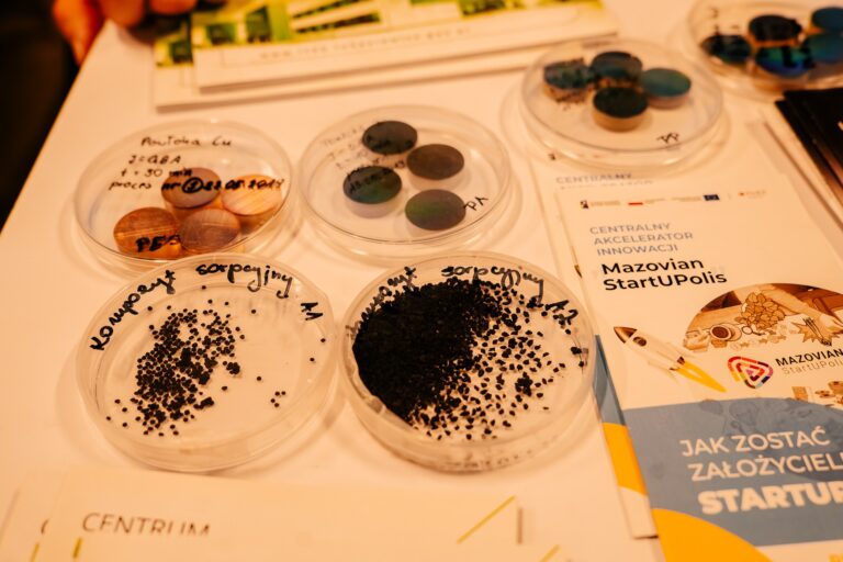 Próbki materiałów badawczych w plastikowych naczyniach laboratoryjnych na stole wystawowym podczas 15. Forum Rozwoju Mazowsza. Obok znajduje się broszura programu „Mazovian StartUPolis – Centralny Akcelerator Innowacji”.