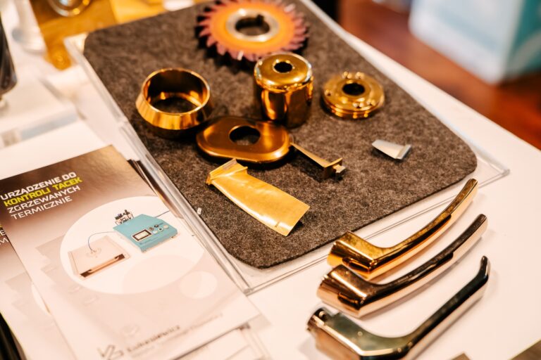 Stoisko Sieci Badawczej Łukasiewicz podczas 15. Forum Rozwoju Mazowsza. Na stole leżą metalowe elementy i próbki powłok technicznych w złotym i srebrnym kolorze, prezentujące możliwości technologiczne instytutu. Obok znajduje się folder informacyjny z logo Łukasiewicza.