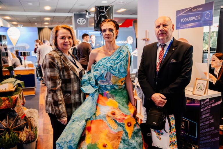 Trzy osoby pozują do zdjęcia podczas 15. Forum Rozwoju Mazowsza. Pośrodku stoi kobieta w artystycznej, kolorowej sukni inspirowanej naturą, a po jej bokach kobieta i mężczyzna w eleganckich strojach biznesowych. W tle widoczne są stoiska wystawców, m.in. z województwa podkarpackiego i łódzkiego.