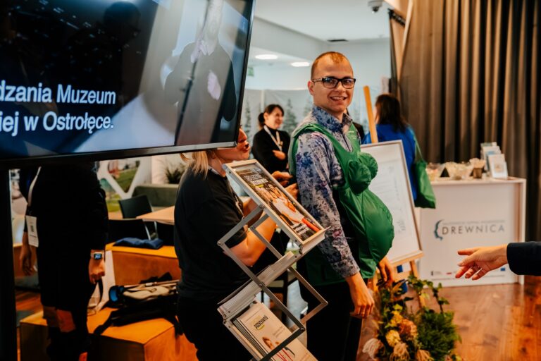 Mężczyzna uśmiecha się, mając na sobie zielony symulator ciąży podczas 15. Forum Rozwoju Mazowsza. Obok niego stoi kobieta z Mazowieckiego Centrum Polityki Społecznej, a w tle widoczne są inne stoiska, w tym Mazowieckiego Centrum Rehabilitacji DREWNICA.