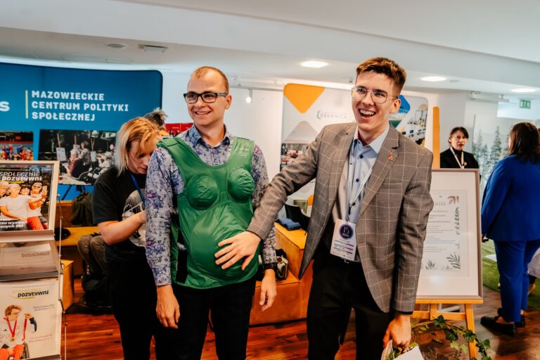 Dwóch mężczyzn uśmiecha się podczas prezentacji na stoisku Mazowieckiego Centrum Polityki Społecznej podczas 15. Forum Rozwoju Mazowsza. Jeden z nich ma na sobie zielony symulator ciąży, a drugi dotyka jego brzucha w żartobliwy sposób. W tle widoczni są inni uczestnicy wydarzenia