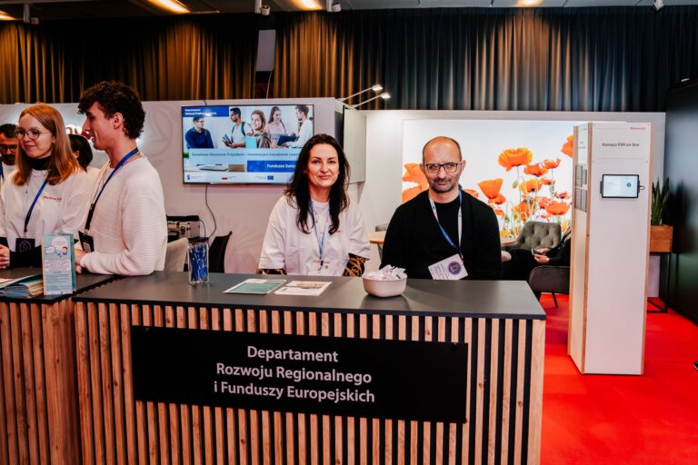 Stoisko Województwa Mazowieckiego – Departamentu Rozwoju Regionalnego i Funduszy Europejskich podczas 15. Forum Rozwoju Mazowsza. Za ladą stoją pracownicy urzędu, a w tle widać ekran z prezentacją i stoisko z tłumaczem PJM on-line.