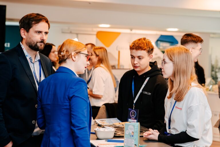 Dwie kobiety stoją przy stoisku prezentującym usługę „Tłumacz PJM on-line” Mazowsza podczas 15. Forum Rozwoju Mazowsza. Na środku znajduje się ekran z interfejsem tłumacza języka migowego, a w tle widać część przestrzeni wystawienniczej.