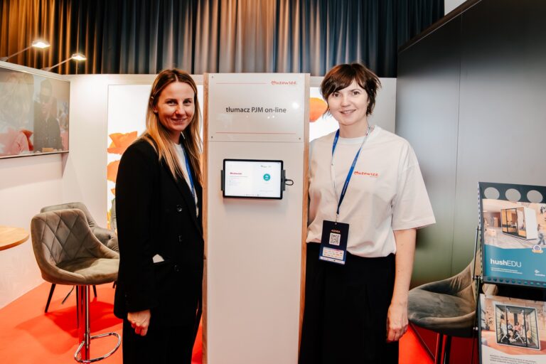 Dwie kobiety stoją przy stoisku prezentującym usługę „Tłumacz PJM on-line” Mazowsza podczas 15. Forum Rozwoju Mazowsza. Na środku znajduje się ekran z interfejsem tłumacza języka migowego, a w tle widać część przestrzeni wystawienniczej