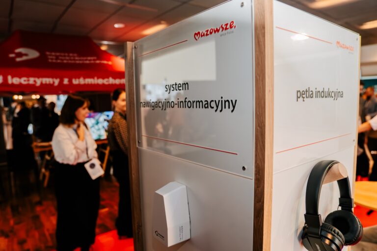 Stoisko promujące system nawigacyjno-informacyjny i pętlę indukcyjną Mazowsza podczas 15. Forum Rozwoju Mazowsza. Na konstrukcji widoczne są słuchawki, a w tle uczestnicy wydarzenia i czerwony namiot szpitala z napisem „Leczymy z uśmiechem”.
