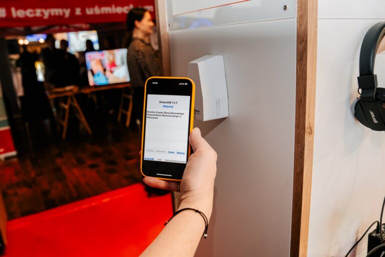 Osoba przykłada smartfon do urządzenia przy stoisku podczas 15. Forum Rozwoju Mazowsza. Na ekranie telefonu widoczny jest komunikat dotyczący stoiska Urzędu Marszałkowskiego Województwa Mazowieckiego w Warszawie. W tle uczestnicy wydarzenia i czerwony baner.