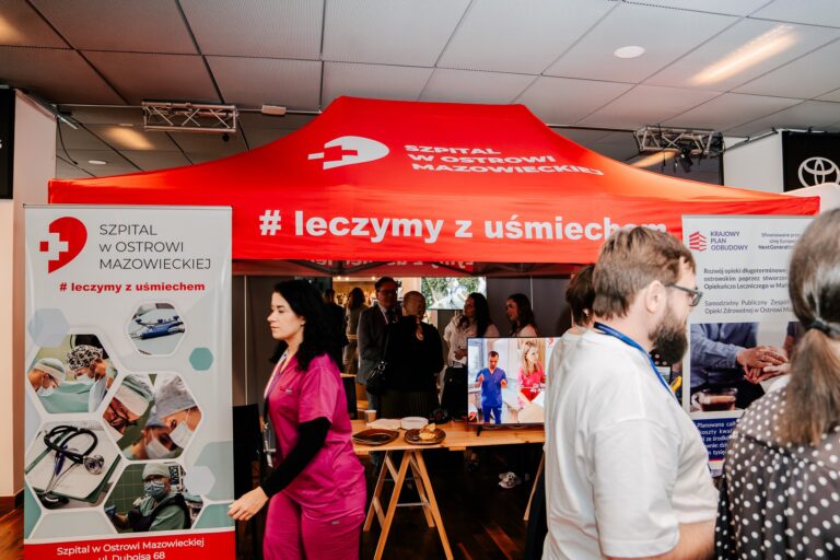 Stoisko Szpitala w Ostrowi Mazowieckiej podczas 15. Forum Rozwoju Mazowsza. Na pierwszym planie widać kobietę w medycznym uniformie, a w tle czerwoną konstrukcję z napisem „Leczymy z uśmiechem” oraz uczestników wydarzenia rozmawiających przy stoisku.