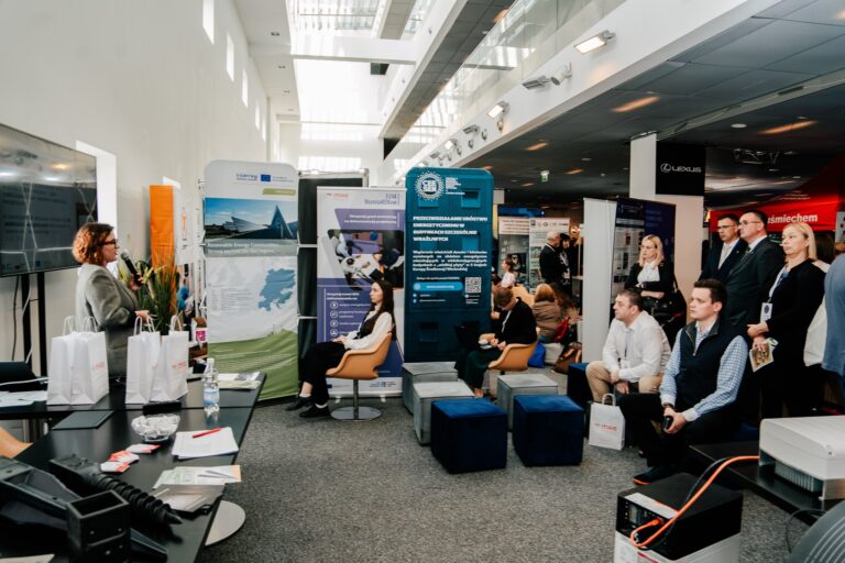 Przedstawicielka Mazowieckiej Agencji Energetycznej prowadzi prezentację podczas 15. Forum Rozwoju Mazowsza. Uczestnicy siedzą i stoją przed stoiskami z banerami dotyczącymi energii odnawialnej i efektywności energetycznej.