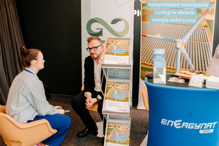 Kobieta i mężczyzna rozmawiają przy stoisku informacyjnym podczas 15. Forum Rozwoju Mazowsza. Na stoliku i stojaku widoczne są broszury oraz materiały promocyjne dotyczące energii i zrównoważonego rozwoju.