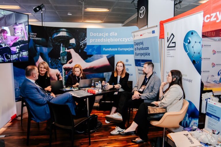 Grupa uczestników 15. Forum Rozwoju Mazowsza prowadzi rozmowę przy stoisku Sieci Badawczej Łukasiewicz. W tle widoczne są banery informacyjne o funduszach europejskich i wsparciu dla przedsiębiorców.